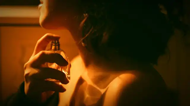 Pessoa aplicando perfume no pescoço com frasco dourado, em luz quente e ambiente escuro