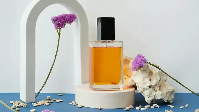 Frasco de perfume âmbar com tampa preta sobre pedestal branco, com flores roxas e fundo azul