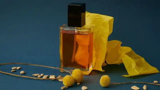 Frasco de perfume âmbar com tampa preta, papel amarelo e flores secas sobre fundo azul-escuro