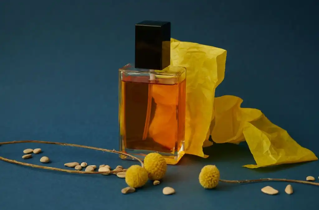 Frasco de perfume âmbar com tampa preta, papel amarelo e flores secas sobre fundo azul-escuro