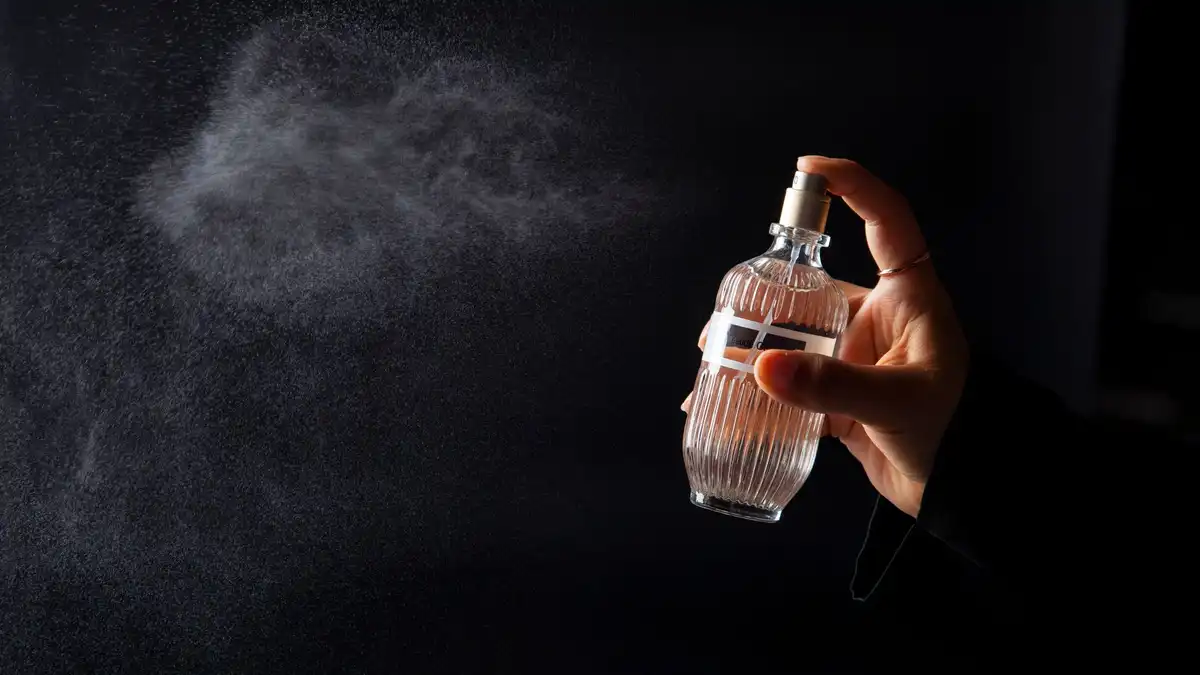 Mão borrifando um frasco de Eau de Parfum, com a névoa da fragrância visível sobre fundo escuro