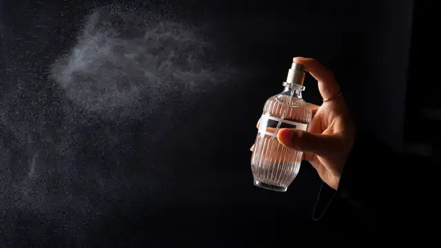 Mão borrifando um frasco de Eau de Parfum, com a névoa da fragrância visível sobre fundo escuro