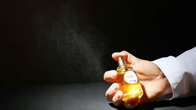 Mão borrifando um frasco de perfume dourado formando uma nuvem de névoa contra fundo escuro