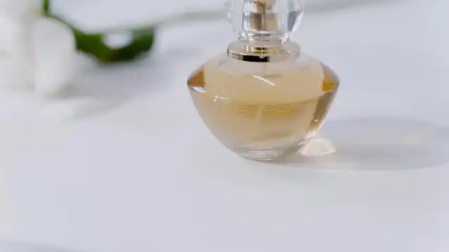 Frasco de perfume feminino dourado com tampa de cristal sobre superfície clara com flores brancas