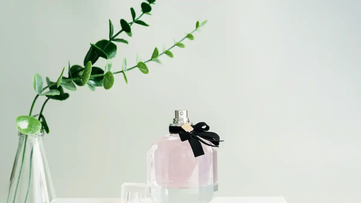 Frasco de perfume rosado com laço preto sobre fundo neutro e ramo de eucalipto