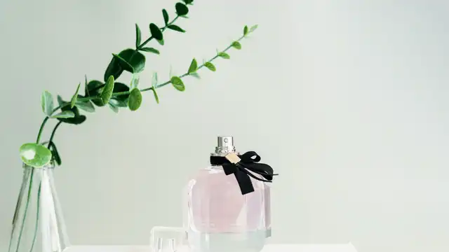 Frasco de perfume rosado com laço preto sobre fundo neutro e ramo de eucalipto