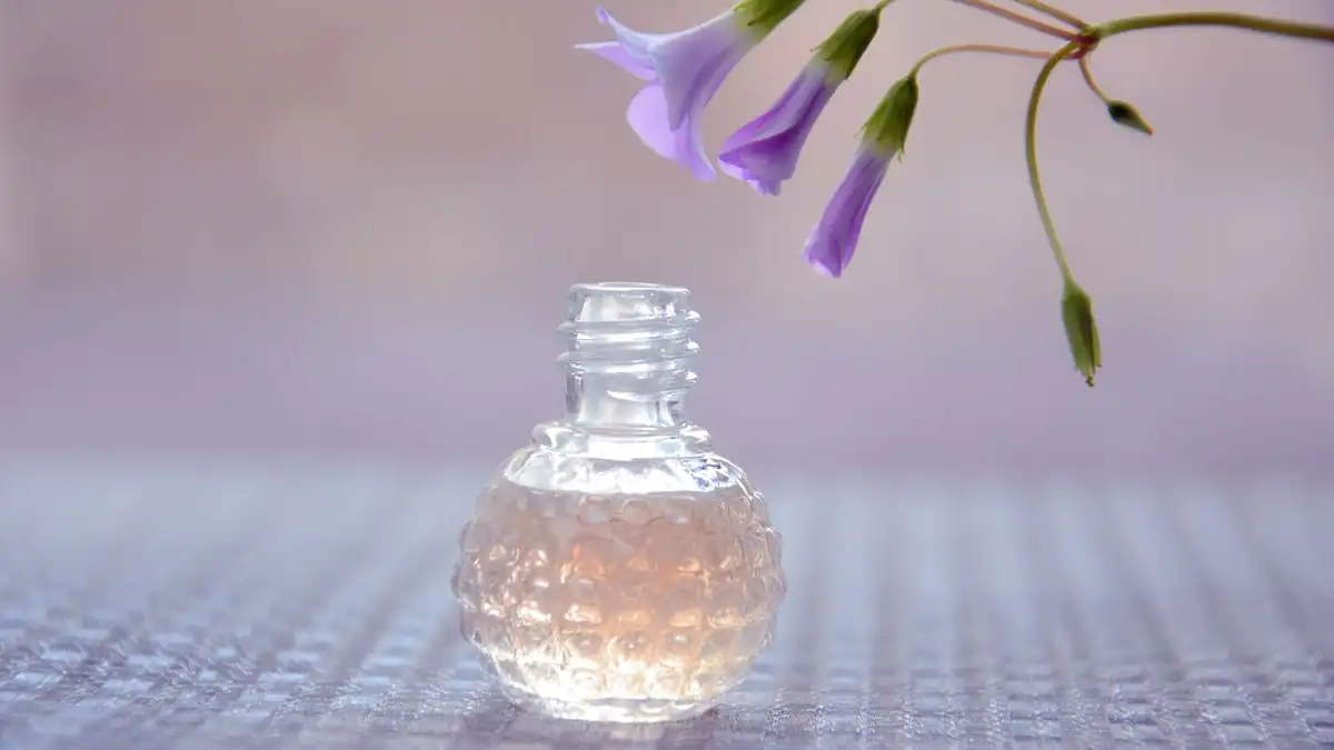 Pequeno frasco de perfume sem rótulo com flor delicada sobre fundo lilás suave