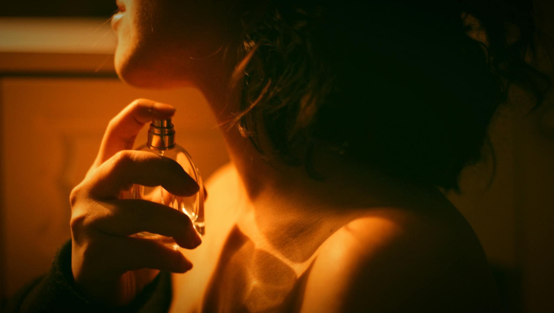 Pessoa aplicando perfume no pescoço com frasco dourado, em luz quente e ambiente escuro