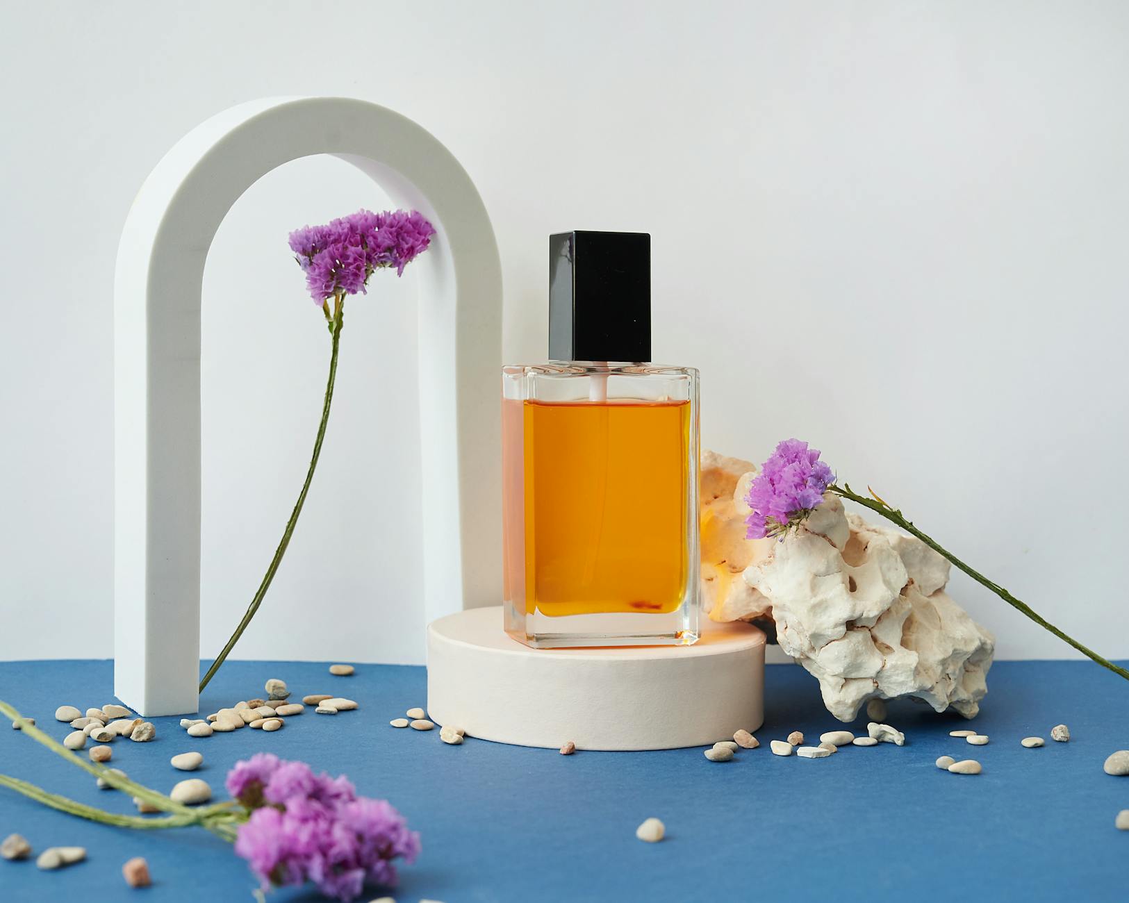 Frasco de perfume âmbar com tampa preta sobre pedestal branco, com flores roxas e fundo azul