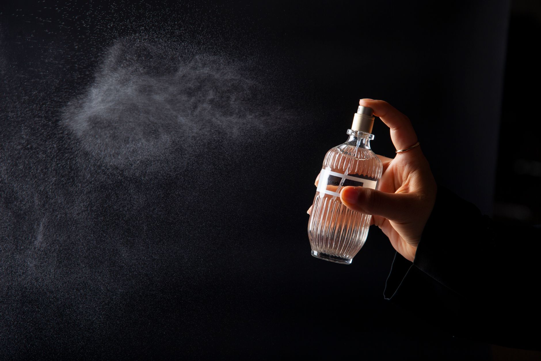 Mão borrifando um frasco de Eau de Parfum, com a névoa da fragrância visível sobre fundo escuro