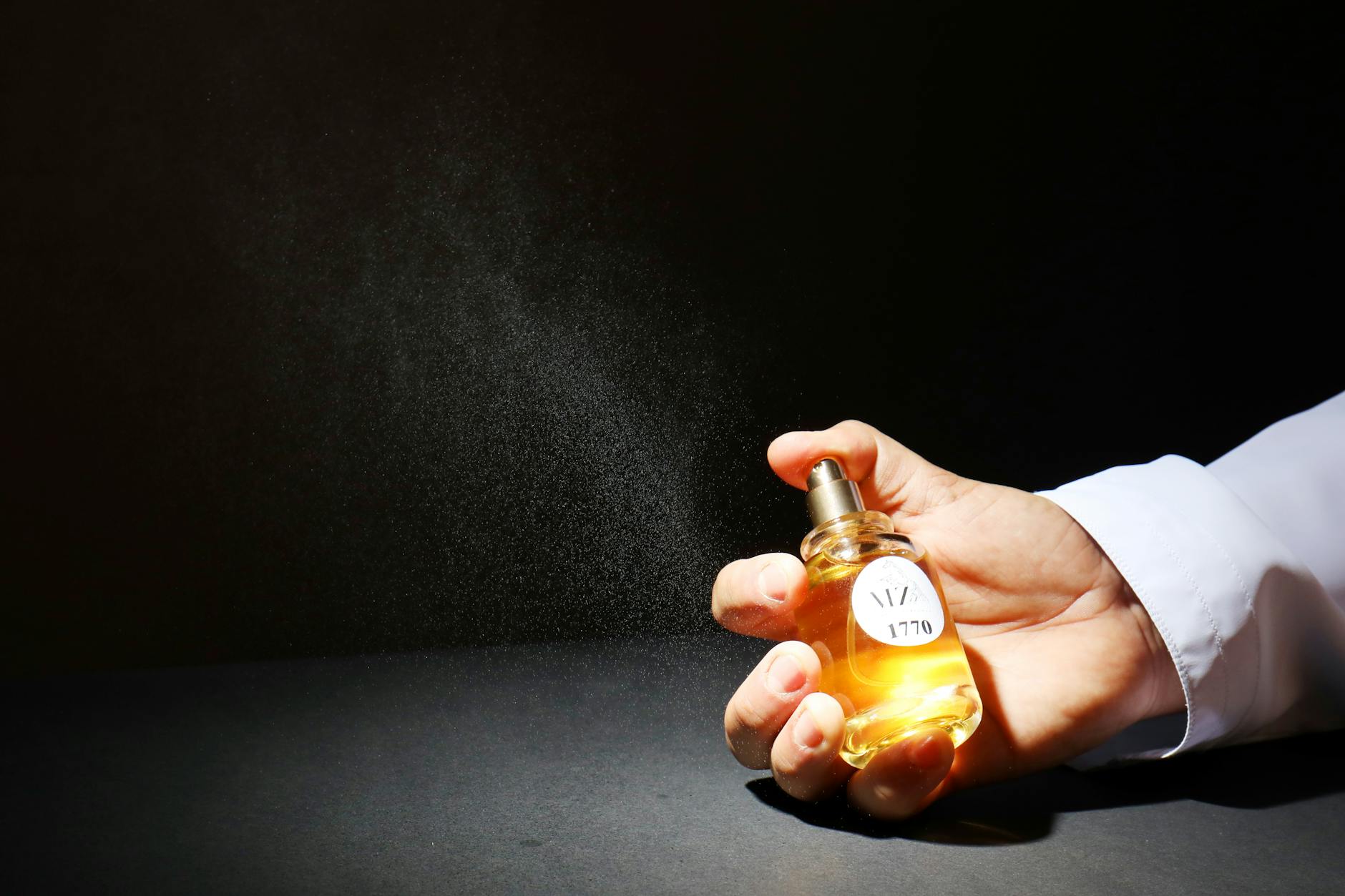 Mão borrifando um frasco de perfume dourado formando uma nuvem de névoa contra fundo escuro